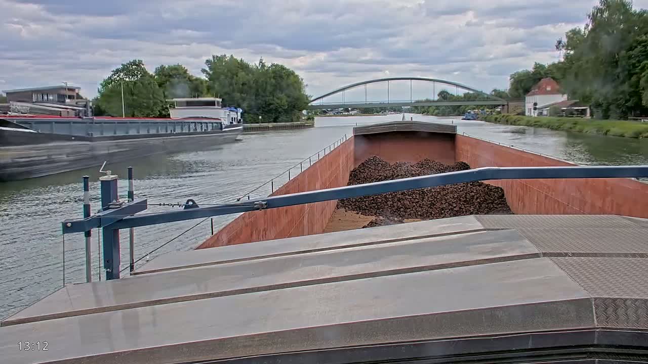 A barge carrying a large pile of dark-colored material travels down a canal under cloudy skies, passing another barge and a bridge.