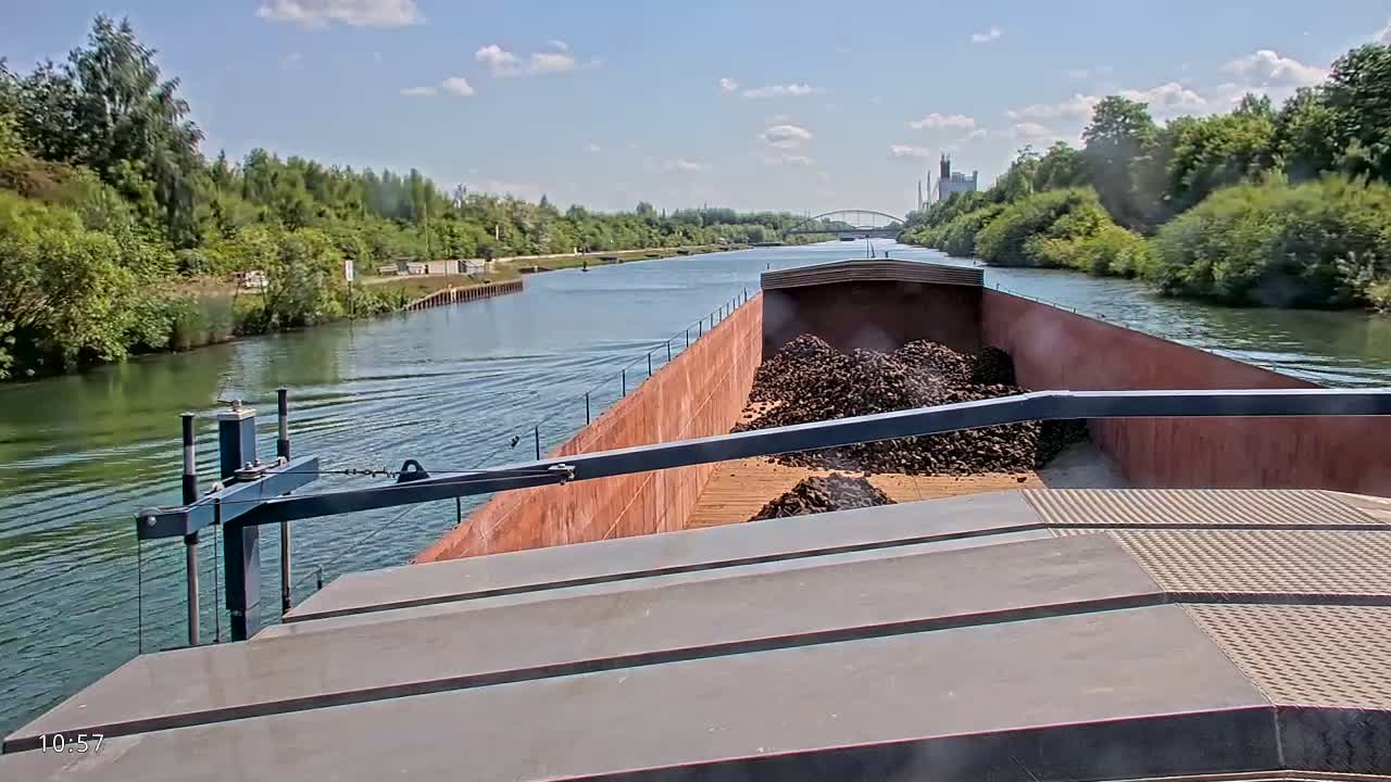A barge carrying a large pile of dark-colored material travels down a canal on a sunny day with partly cloudy skies.
