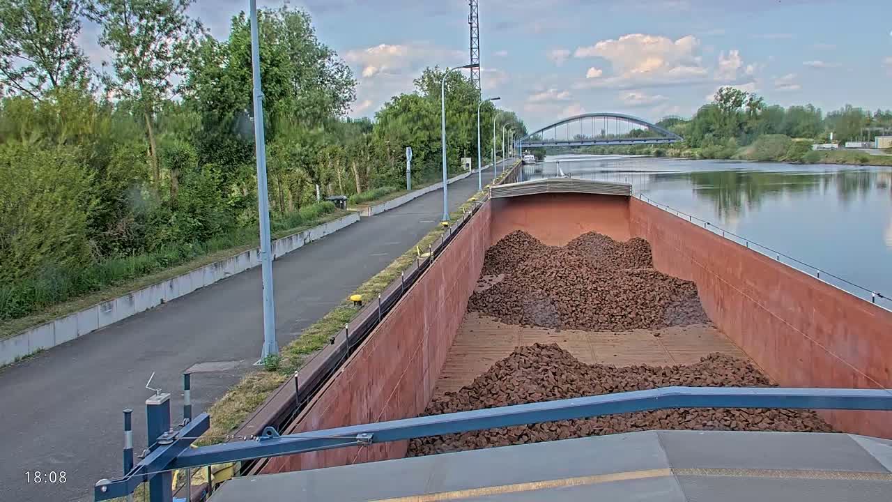 A barge filled with dark brown rocks sits on a calm river under a partly cloudy sky, next to a paved path and lush green vegetation.