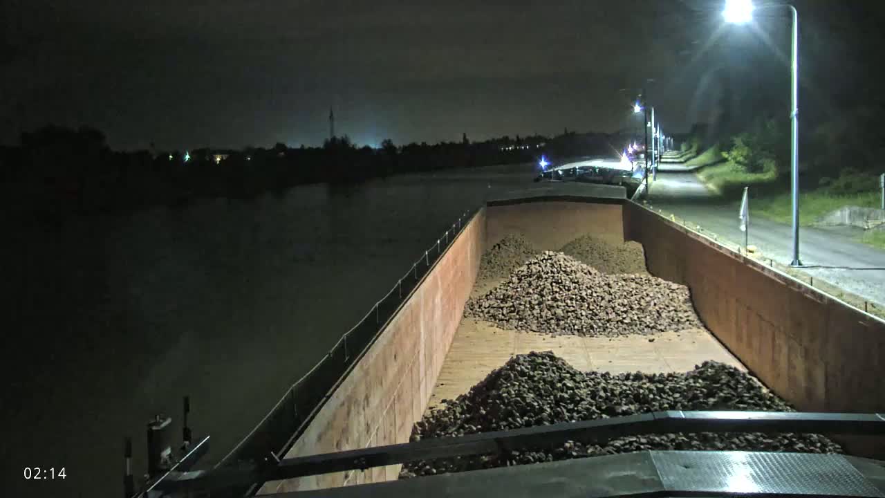 At night, a barge filled with piles of dark-colored rocks is illuminated by streetlights on a nearby roadway next to a body of dark water.