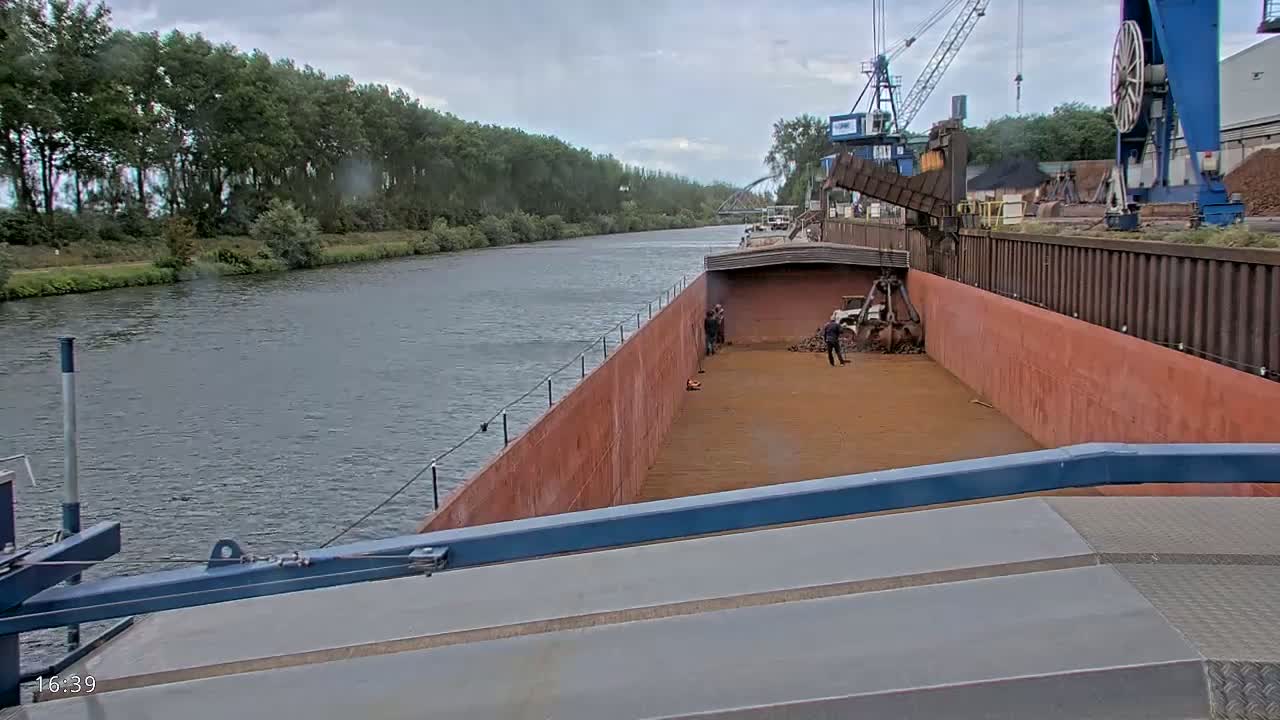 A barge being loaded with reddish-brown material on a cloudy day alongside a canal lined with trees.