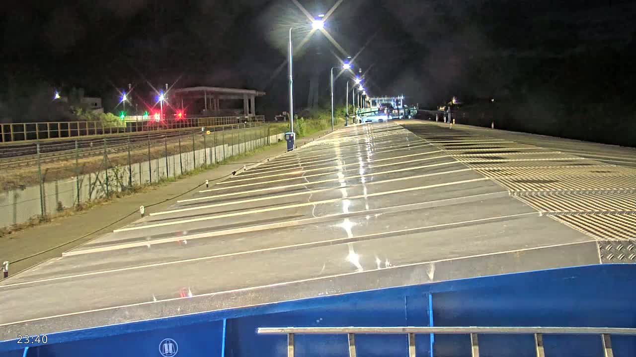 A wet, flat, metal surface stretches toward a nighttime scene of lit train tracks and a fence.