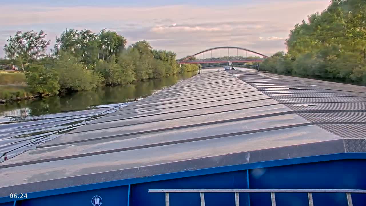 The view from a barge shows a calm river with lush green trees lining the banks, a distant arched bridge, and a partly cloudy sky.