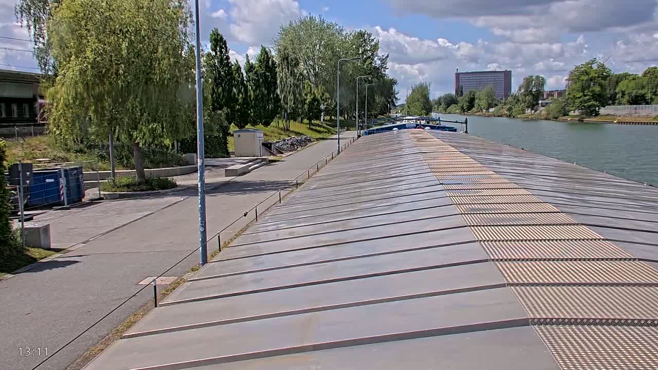 A view from atop a barge on a canal, under partly cloudy skies, shows a paved pathway and lush green trees along the canal bank.