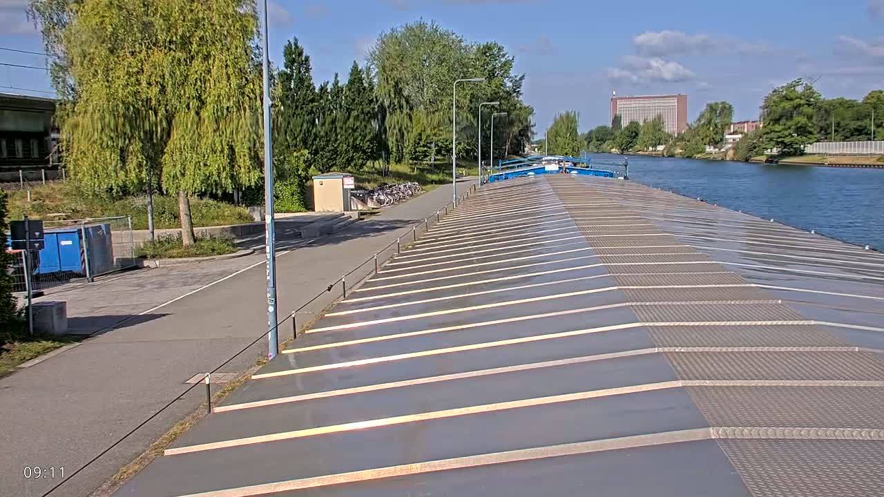 A barge’s flat, gray roof stretches toward a canal under a partly sunny sky, with a paved path and trees lining the waterway.