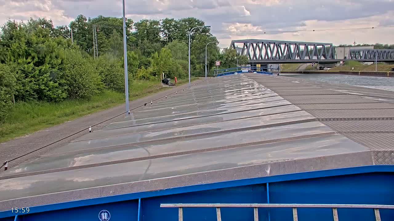 The deck of a barge extends to a canal under a cloudy sky, with a bridge in the distance and lush green vegetation along the canal bank.