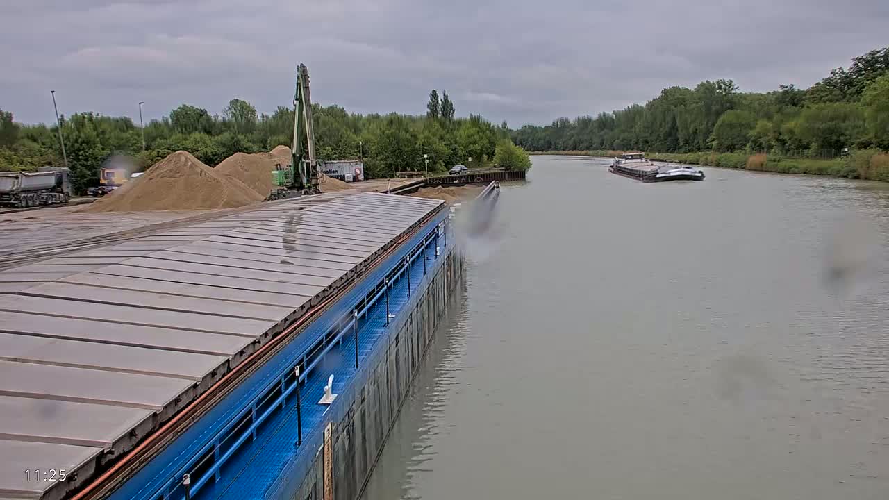 A blue barge is being loaded with sand from a crane on a cloudy day next to a waterway with another barge visible in the distance.