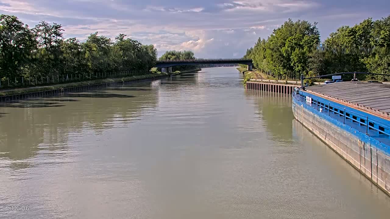 A blue barge is moored alongside a calm, wide canal under a partly cloudy sky, with a bridge visible in the distance and lush green trees lining the banks.