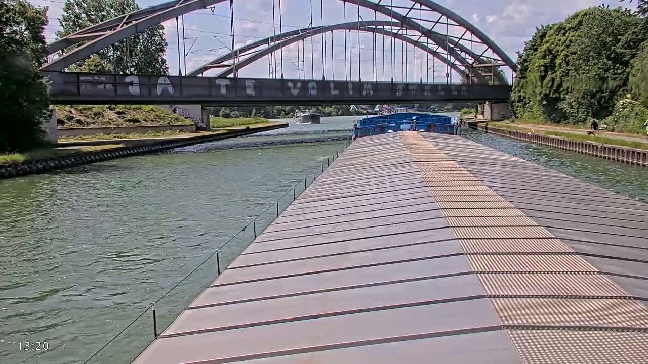 A barge travels on a canal under a large arched bridge on a sunny day.