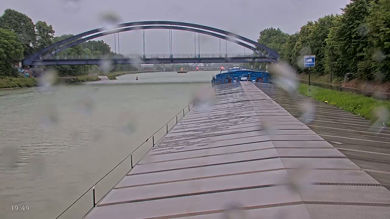 A barge travels on a canal under a steel arch bridge during a rainy day.