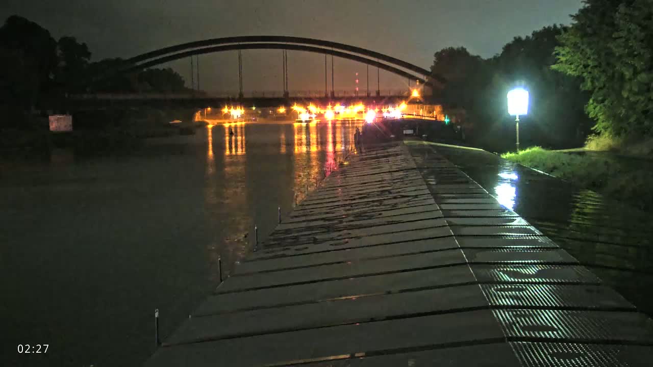 A rain-slicked dock stretches towards a bridge lit by traffic at night, reflecting the lights in the calm water.