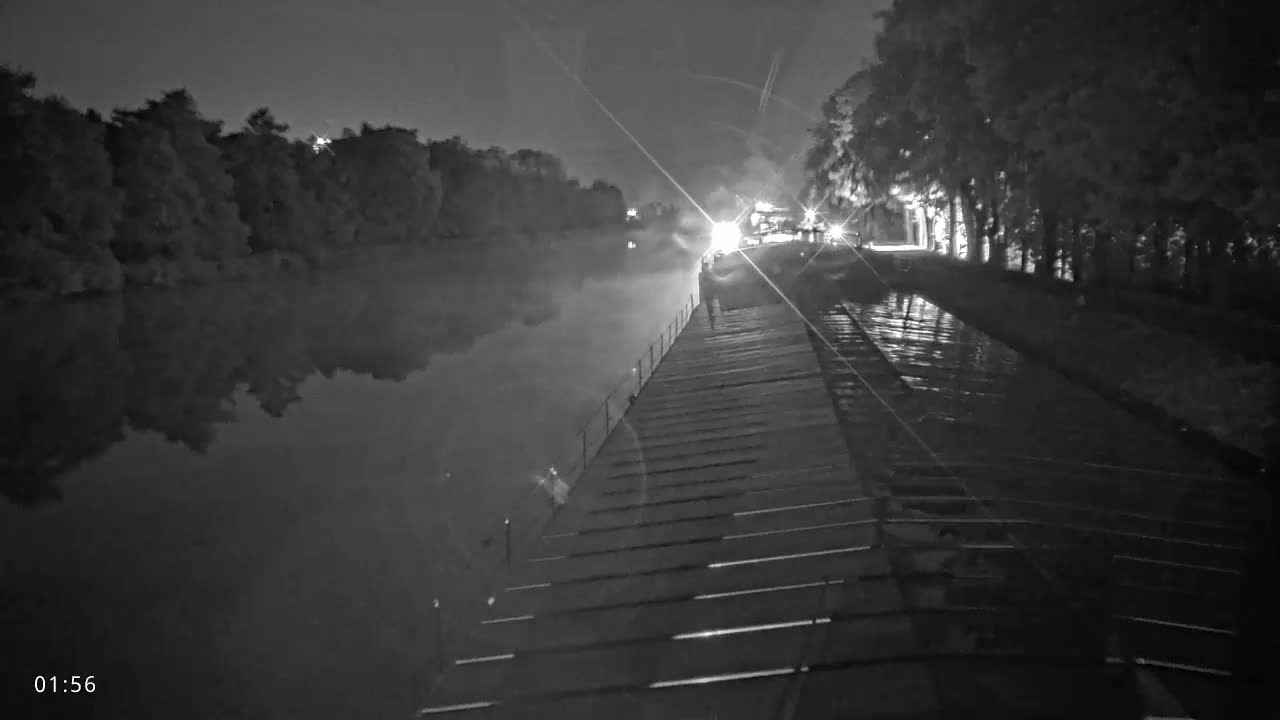 A nighttime view of a long, wooden walkway extending over a calm body of water, lined with trees on either side under a dark sky.