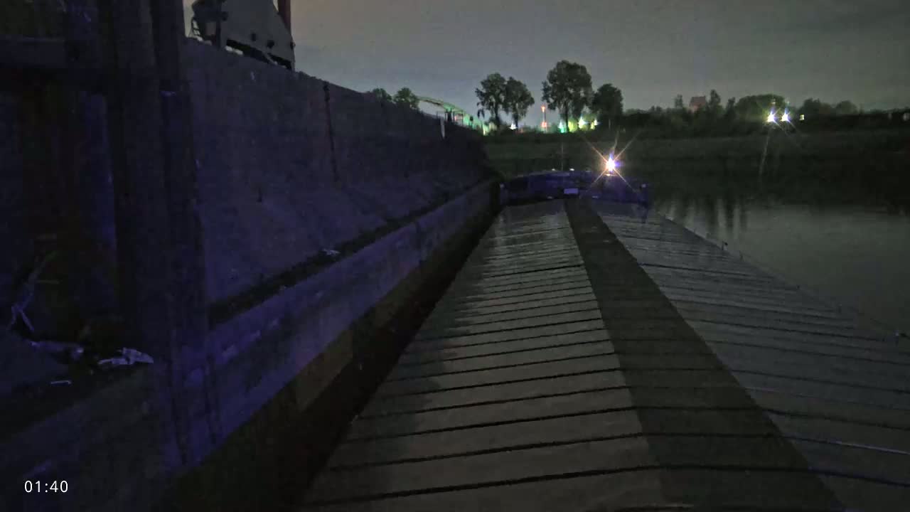 A wet, wooden deck stretches from the camera toward a dark barge on a calm river at night.
