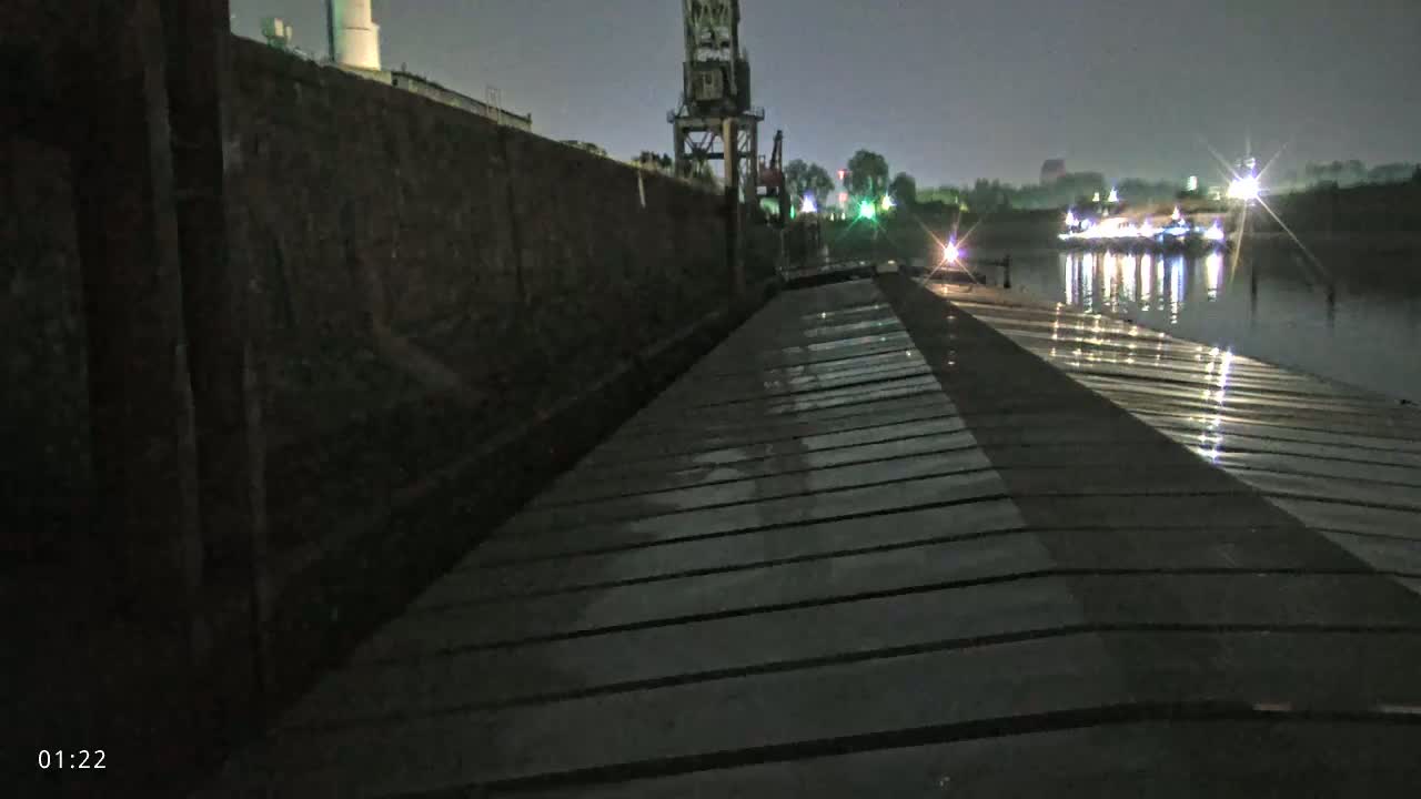 A wet, wooden dock stretches toward a dark waterway at night, alongside a tall, dark wall and distant lights reflecting on the water.