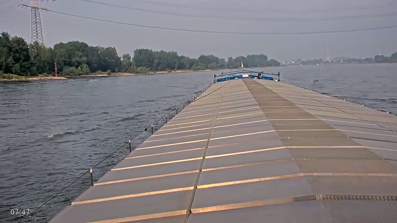 A barge travels down a river under an overcast sky, leaving a small wake in the water.