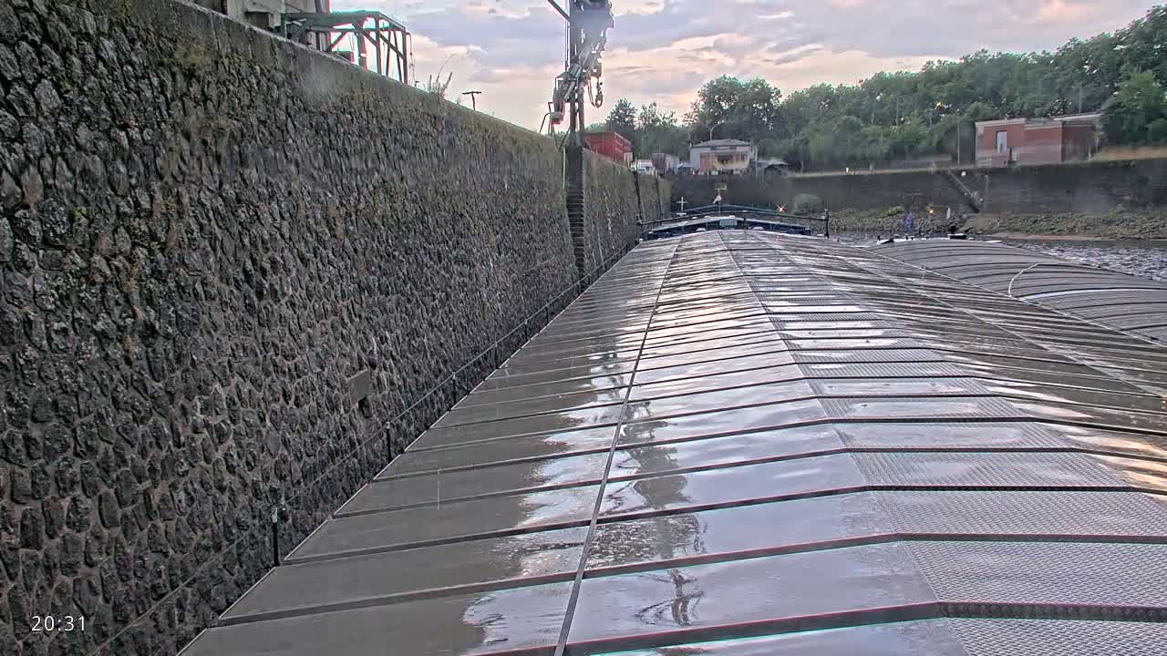 A wet, metal barge deck stretches toward a high stone wall under a cloudy sky.