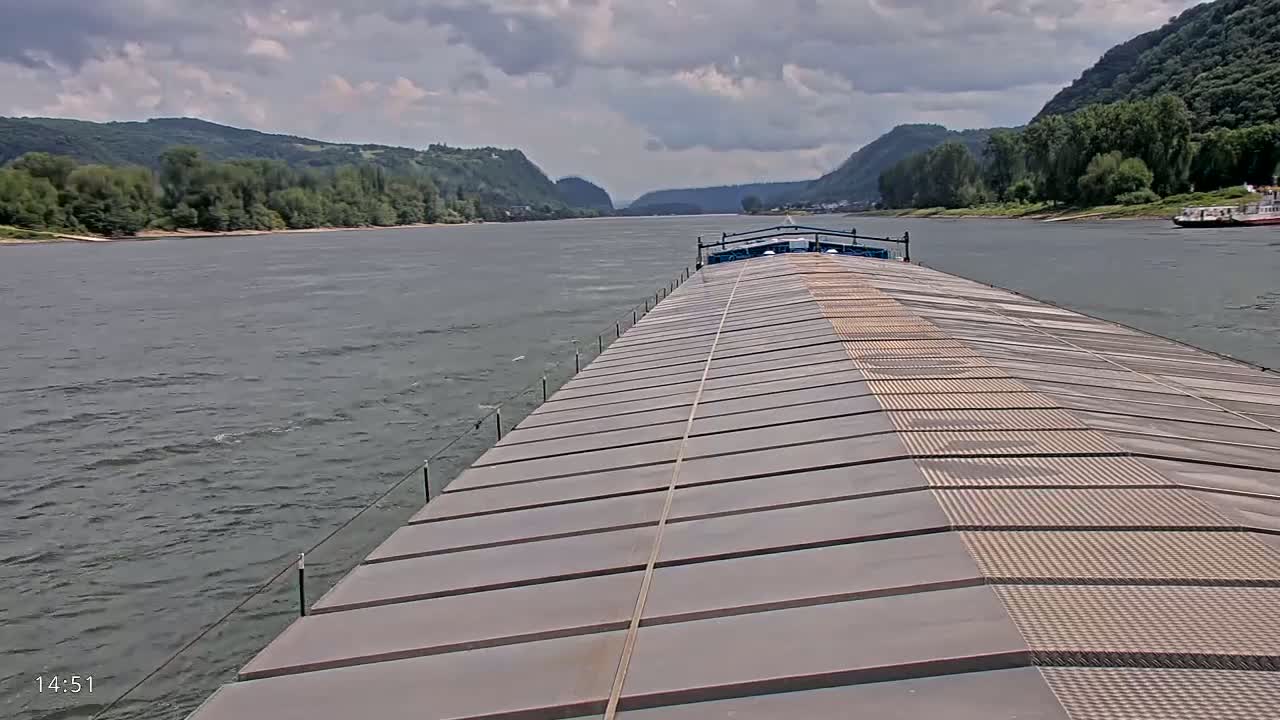 A barge travels down a wide river on a partly cloudy day, with hills and trees visible on either bank.