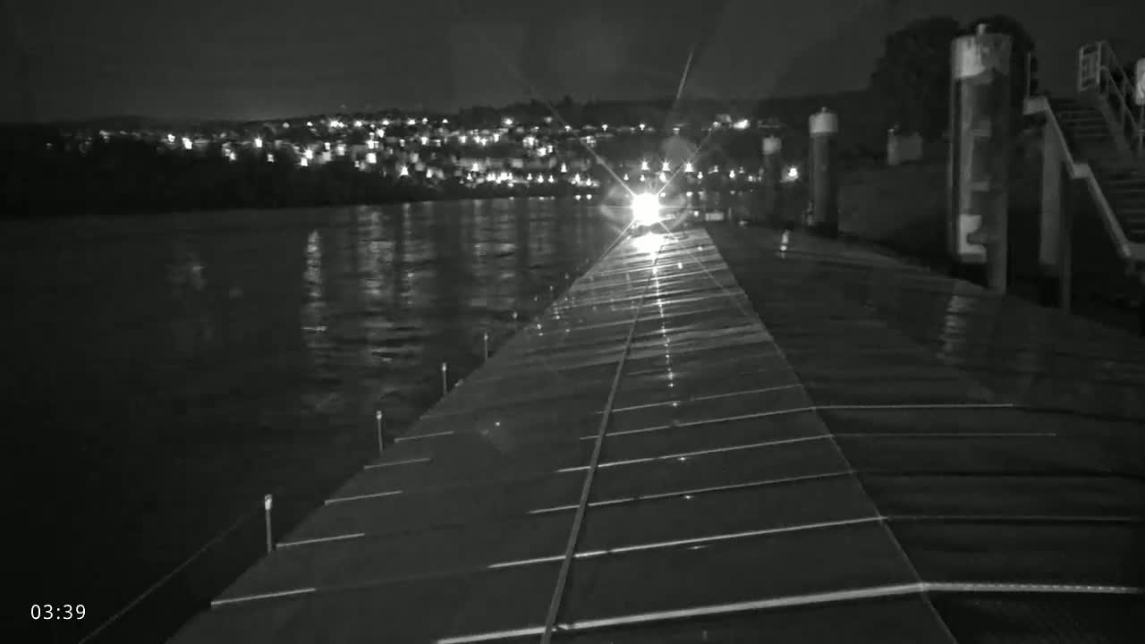 A nighttime view from a vessel's deck shows a dark, wet surface extending toward a distant, lit shoreline across a dark body of water.