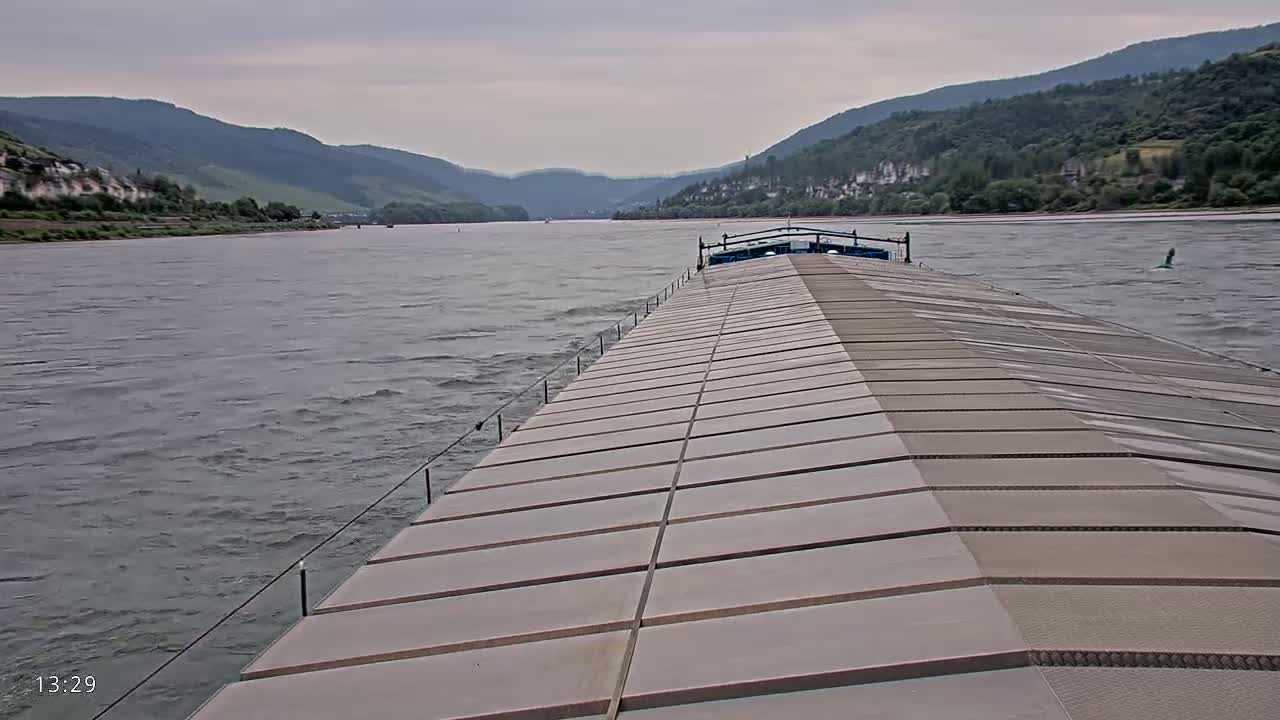 A barge travels down a river under an overcast sky, past hillsides covered in greenery.