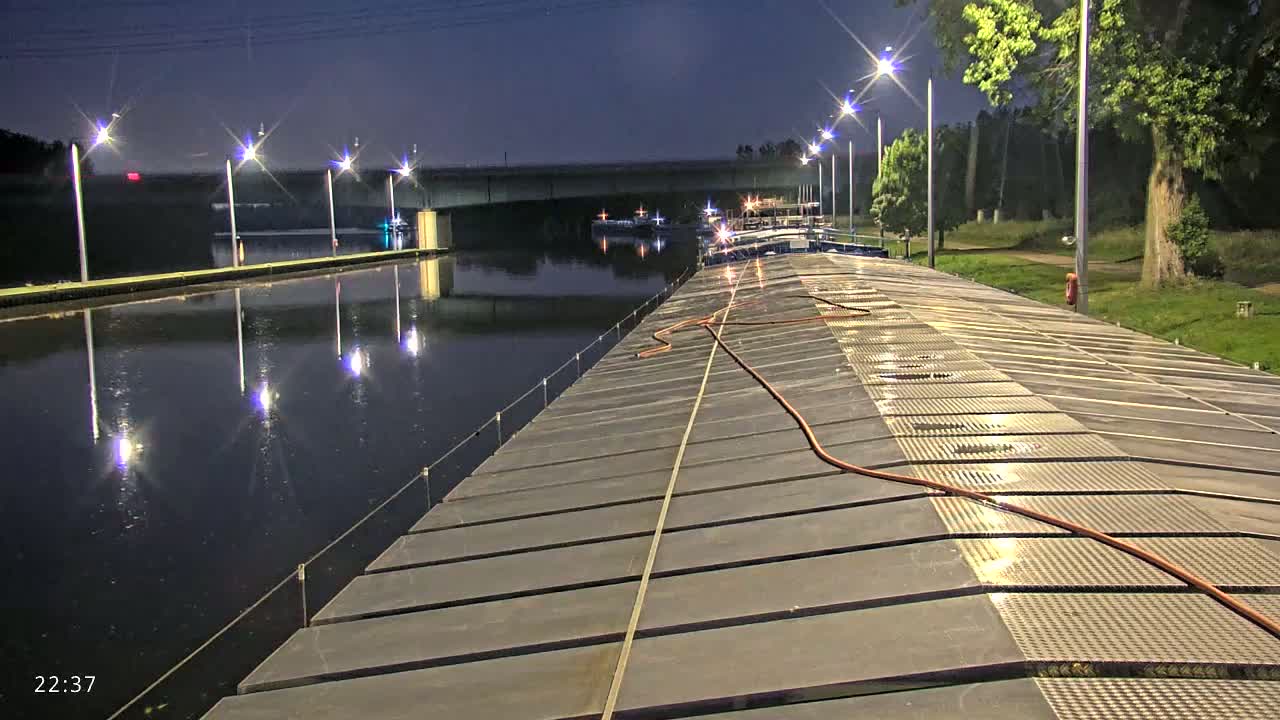 A nighttime view from atop a barge on a calm canal, under a bridge with lights illuminating the water and surrounding dock.