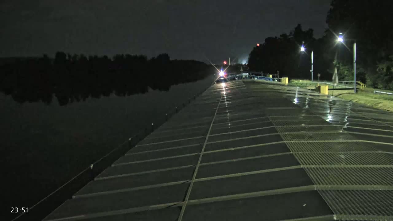 A wet, dark paved area stretches toward a body of water at night, lit by several streetlights.