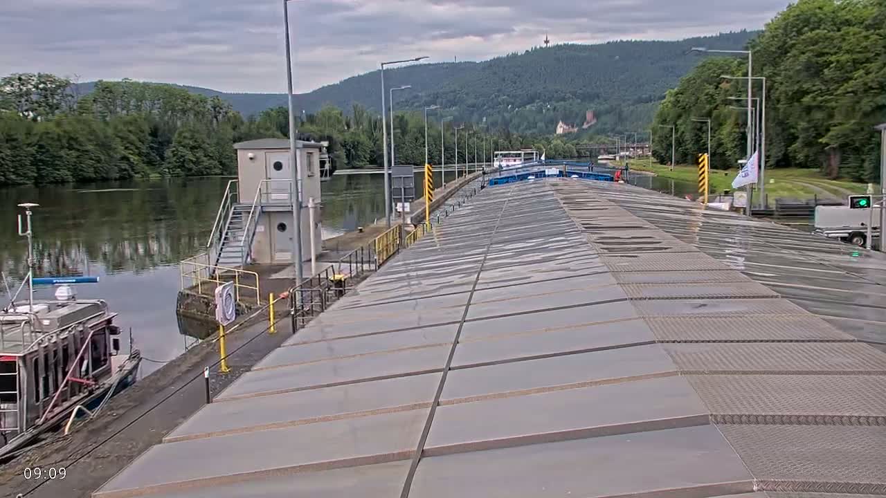 A boat is moored at a lock on a river under a cloudy sky, with hills and trees in the background.