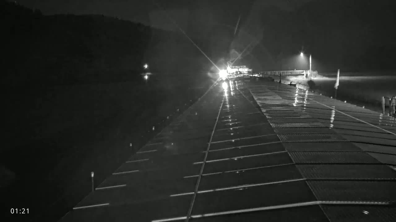 A wet, gridded dock at night is illuminated by headlights approaching from a distance, with a dark shoreline visible in the background.