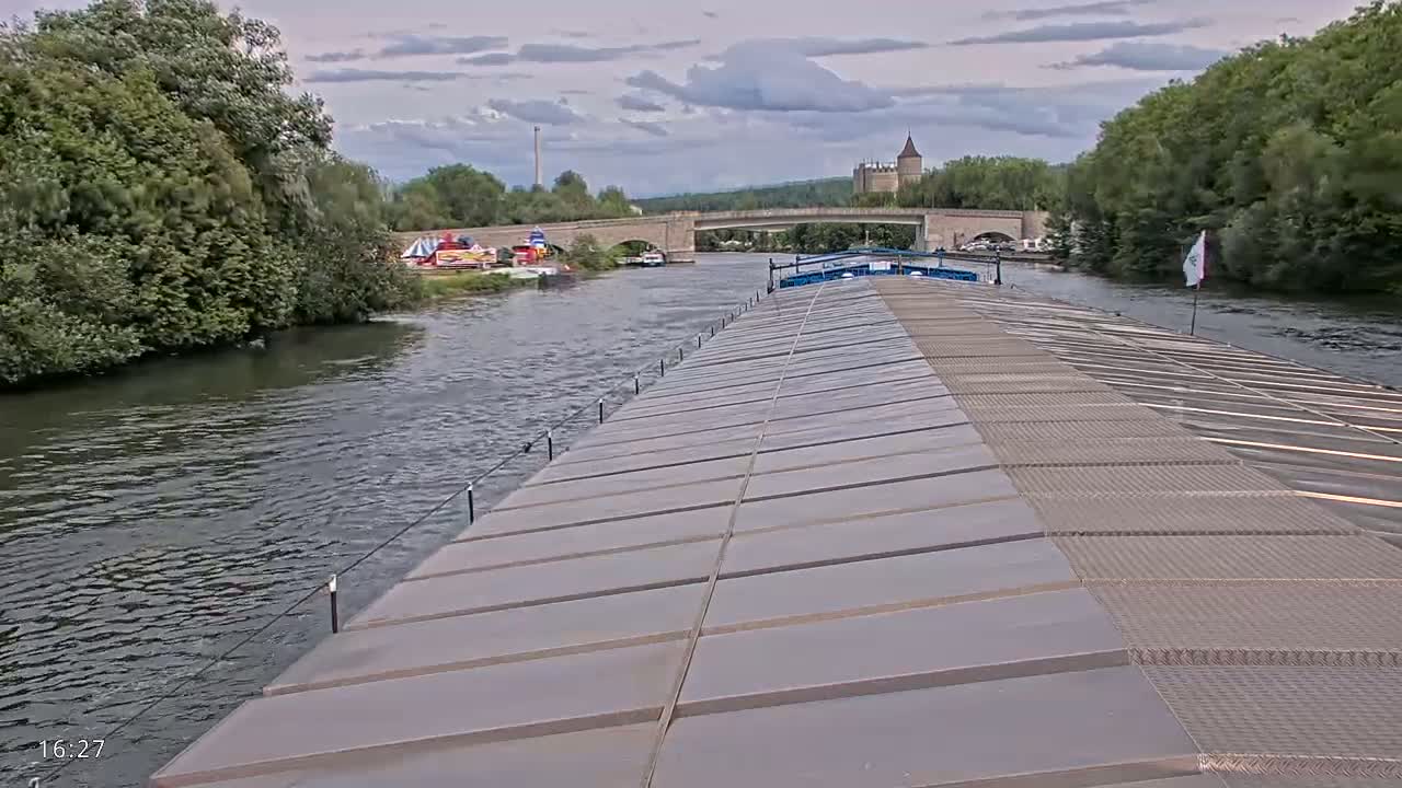 From the deck of a barge, a river scene unfolds under a cloudy sky, showing a stone bridge, a castle in the distance, and lush green vegetation lining the waterway.