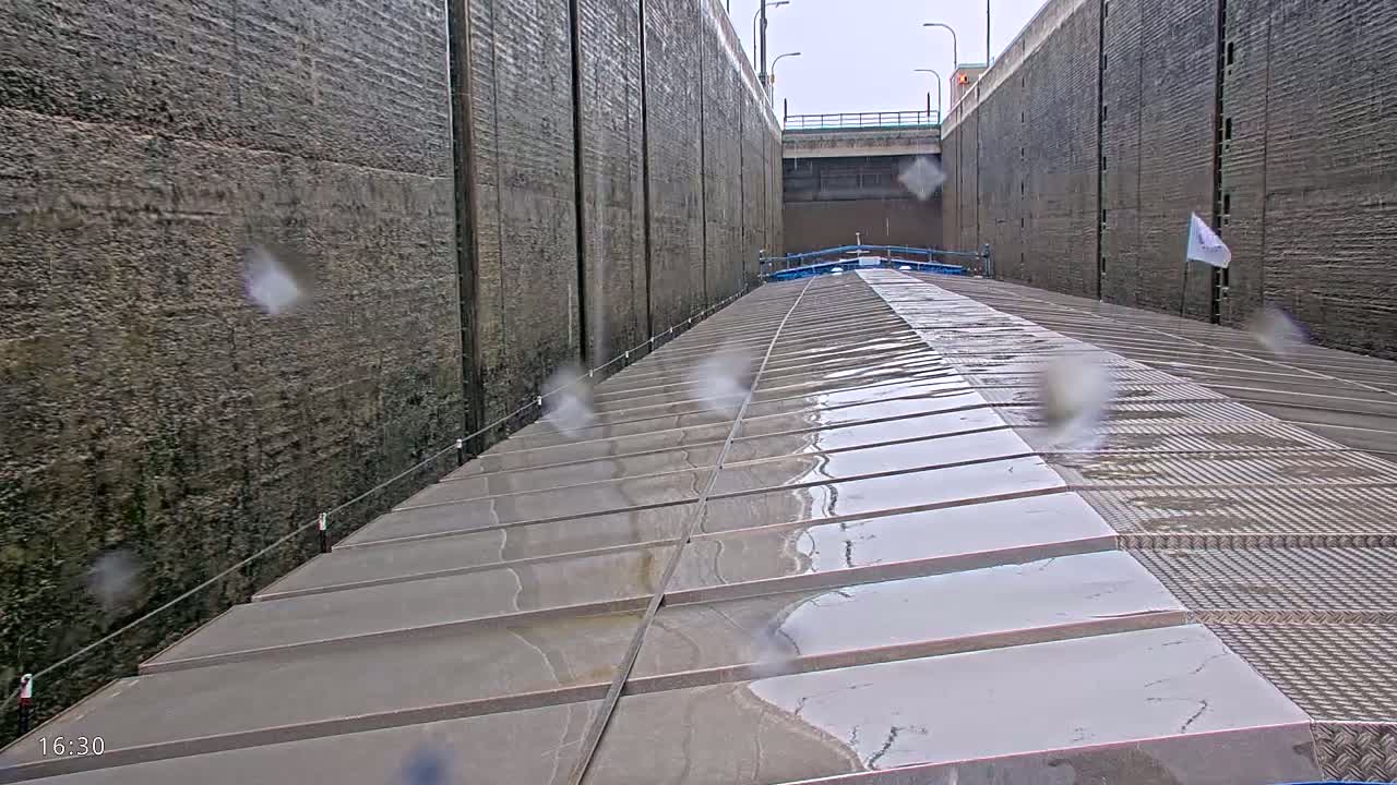 A wet, gray barge deck stretches toward the end of a lock, between tall, gray concrete walls in rainy weather.