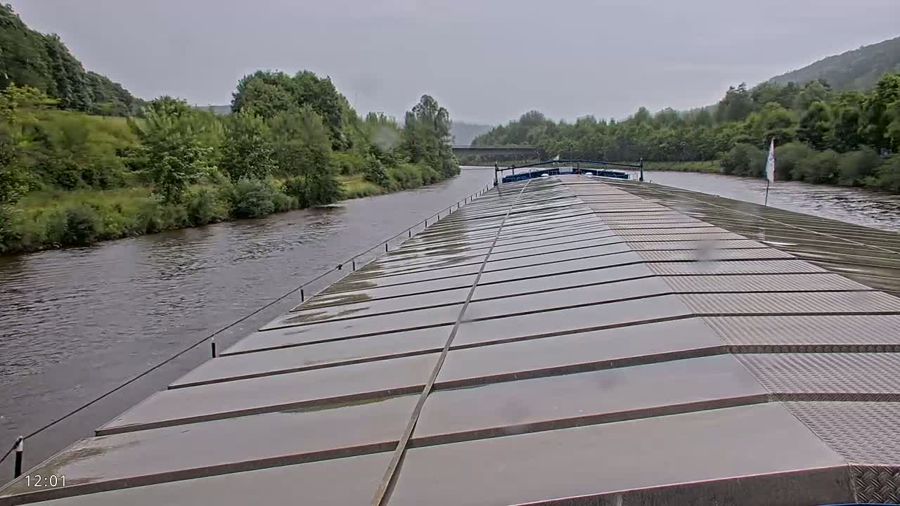 A rain-slicked barge deck stretches to a river bordered by lush green banks and hills under an overcast sky.