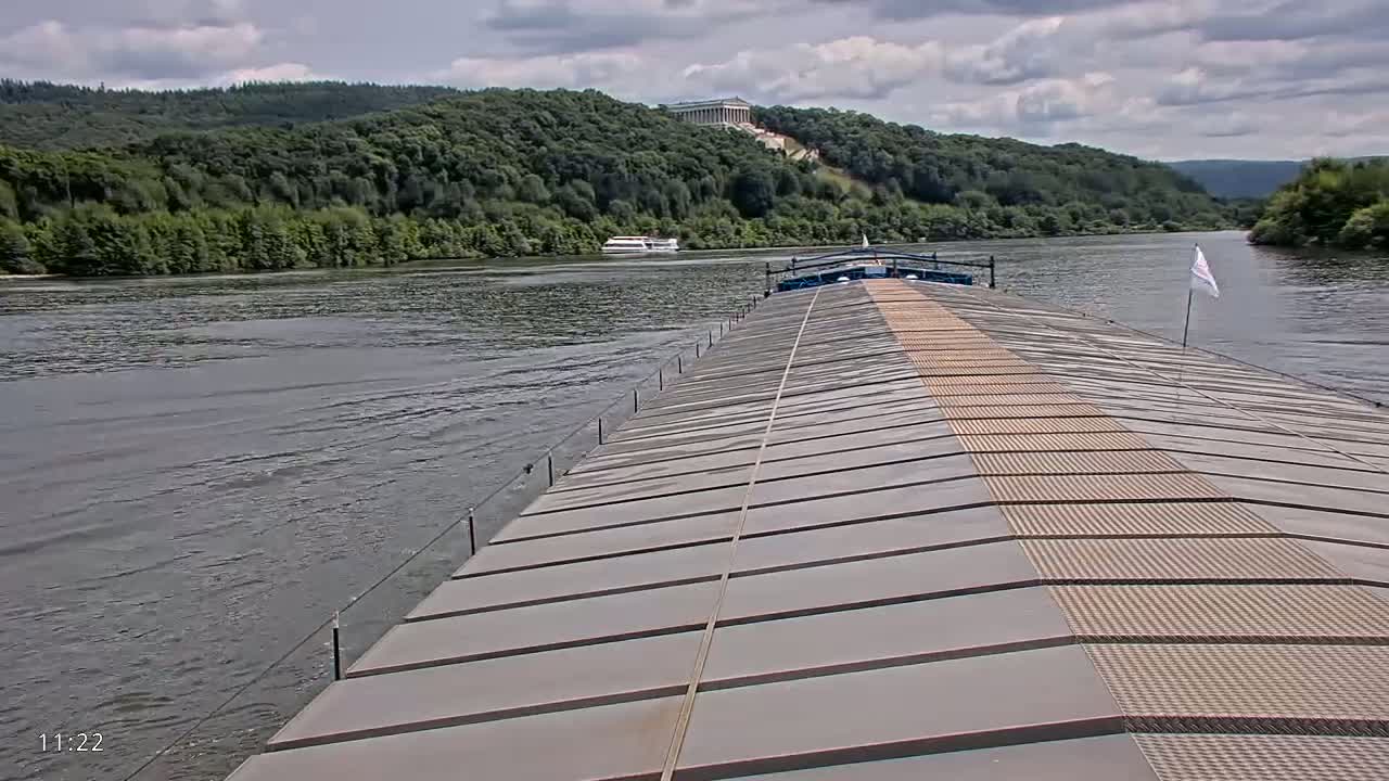 A barge's deck stretches toward a river under a partly cloudy sky, with lush green hills and a distant building visible in the background.