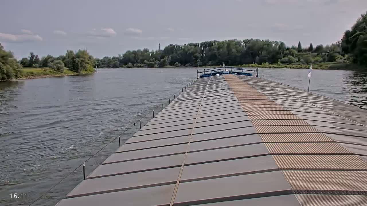 The deck of a barge on a river under partly cloudy skies, with green vegetation visible on both banks.