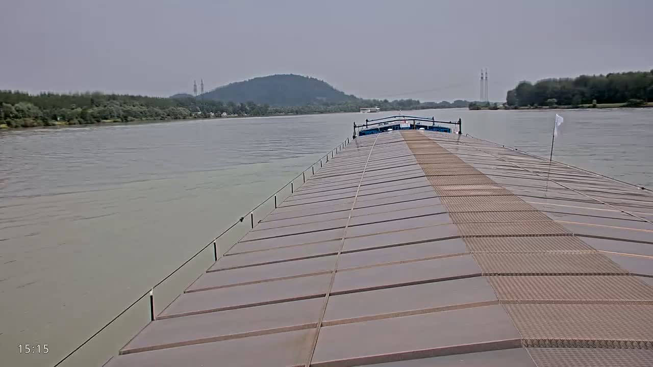A barge's deck stretches toward a river under an overcast sky, with distant hills and trees lining the riverbanks.