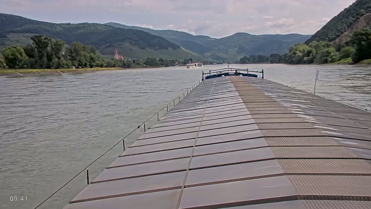 A barge travels down a river under a cloudy sky, with hills and vegetation visible in the distance.