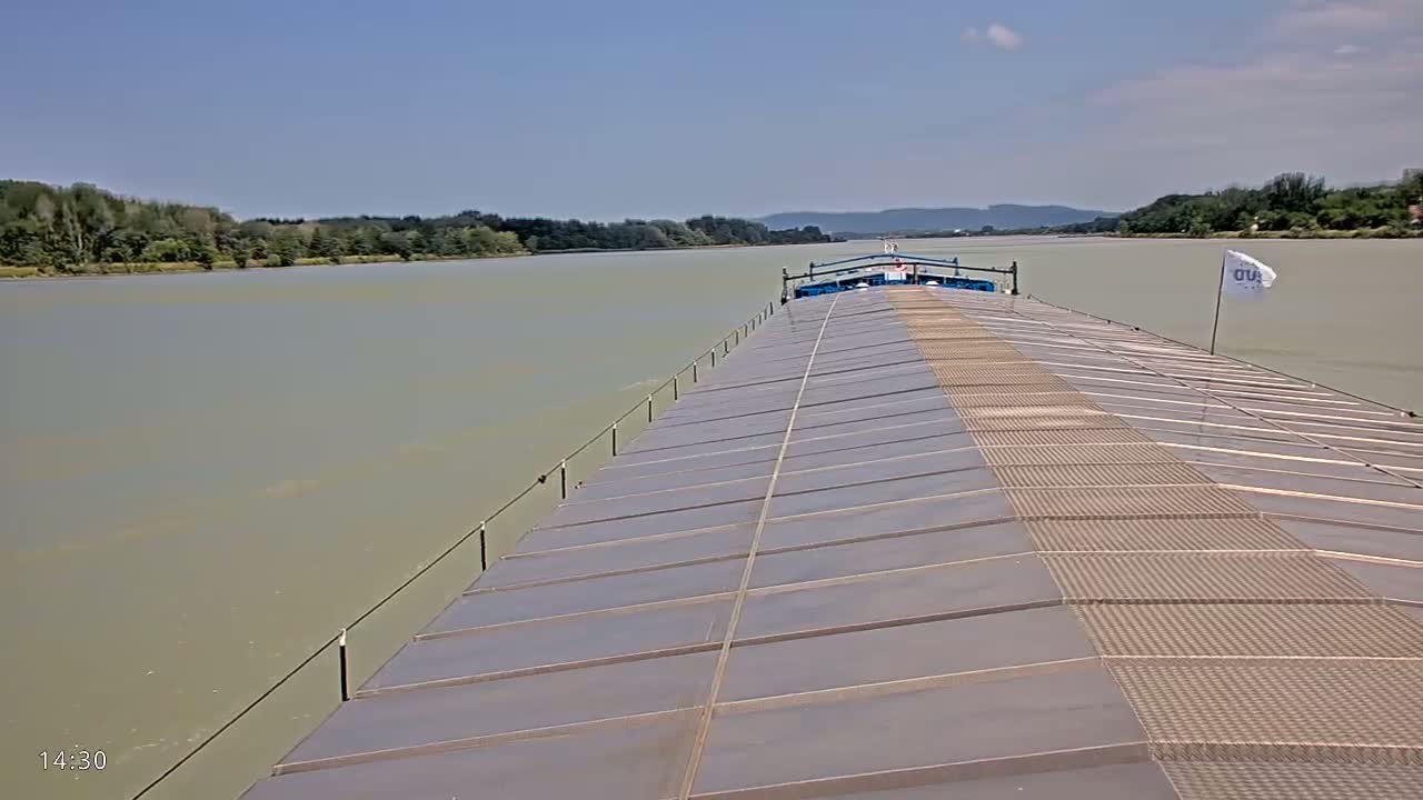 A barge travels down a wide, calm river on a sunny day, with green vegetation visible on either bank.