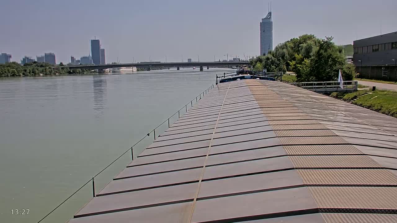 A view from atop a barge on a calm river, showing a cityscape with skyscrapers and a bridge in the distance under a clear sky.