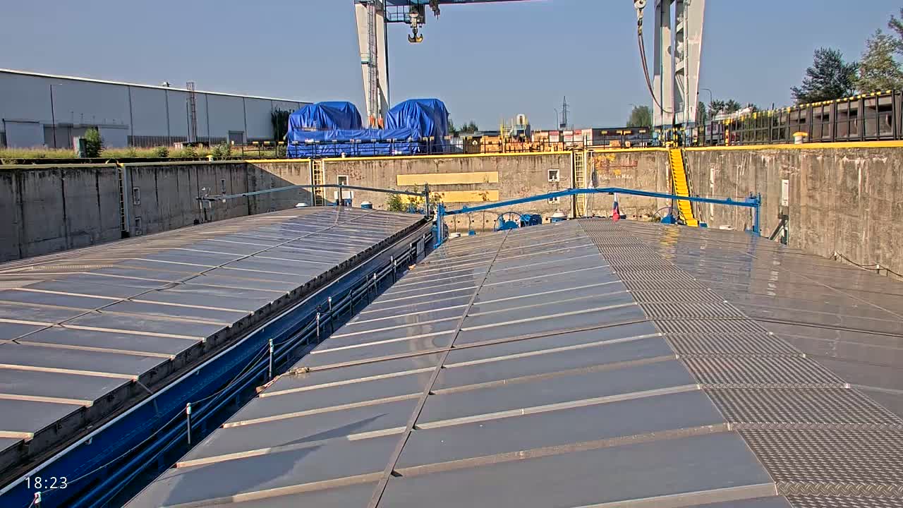 A large, flat, gray surface with a metal walkway down the center is situated within a concrete dock on a sunny day, with a crane and covered cargo visible in the background.