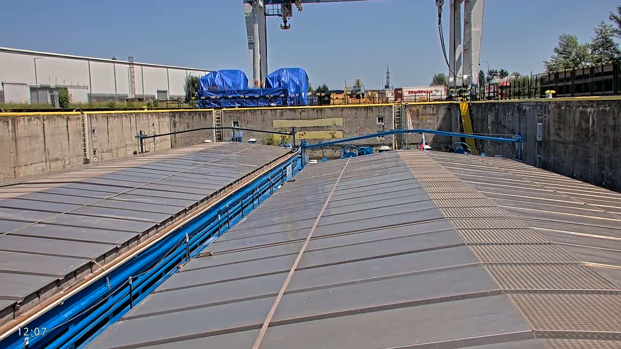 A long, gray, flat-topped barge is situated in a lock, under a sunny sky.