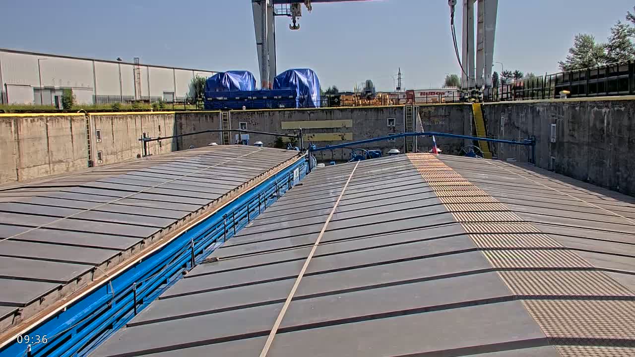 A view from atop a barge shows two gray, slatted decks under a sunny sky, within a concrete dock; a large crane is visible in the background.