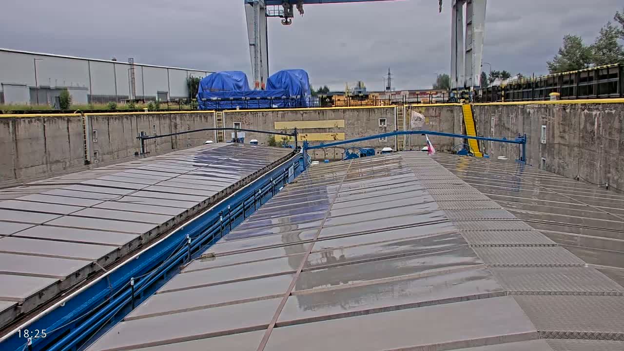 A wet, gray, flat-topped barge sits in a concrete dock under an overcast sky, with industrial buildings and cranes visible in the background.