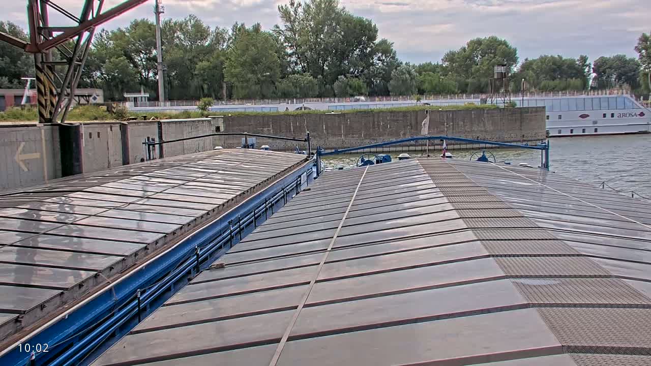 The deck of a barge, with a mostly overcast sky, overlooks a river where another boat is moored along a tree-lined bank.