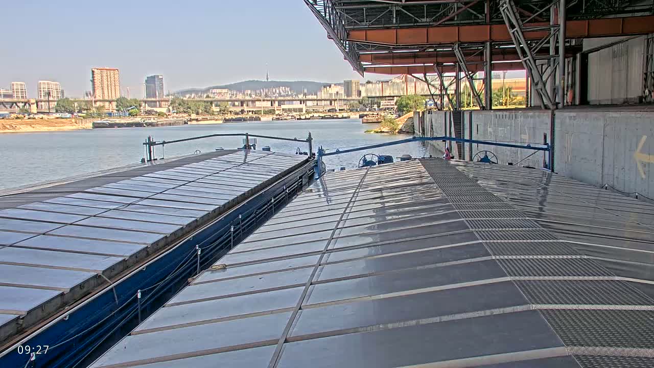 Two barges are docked at a port on a sunny day, with a city skyline visible in the background.