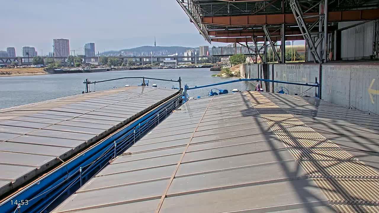 The deck of a barge is shown, with a cityscape and river visible in the background under a sunny sky.