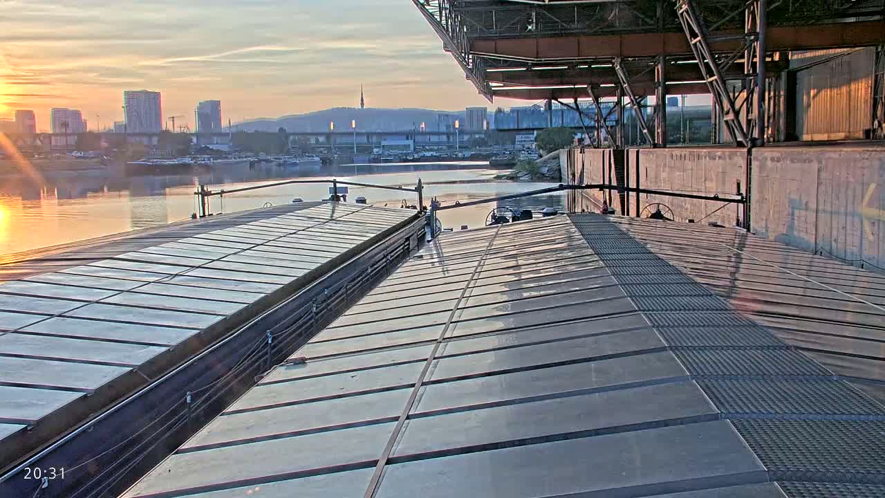 Two barges are docked at sunrise next to a large industrial structure, with a city skyline visible in the background.