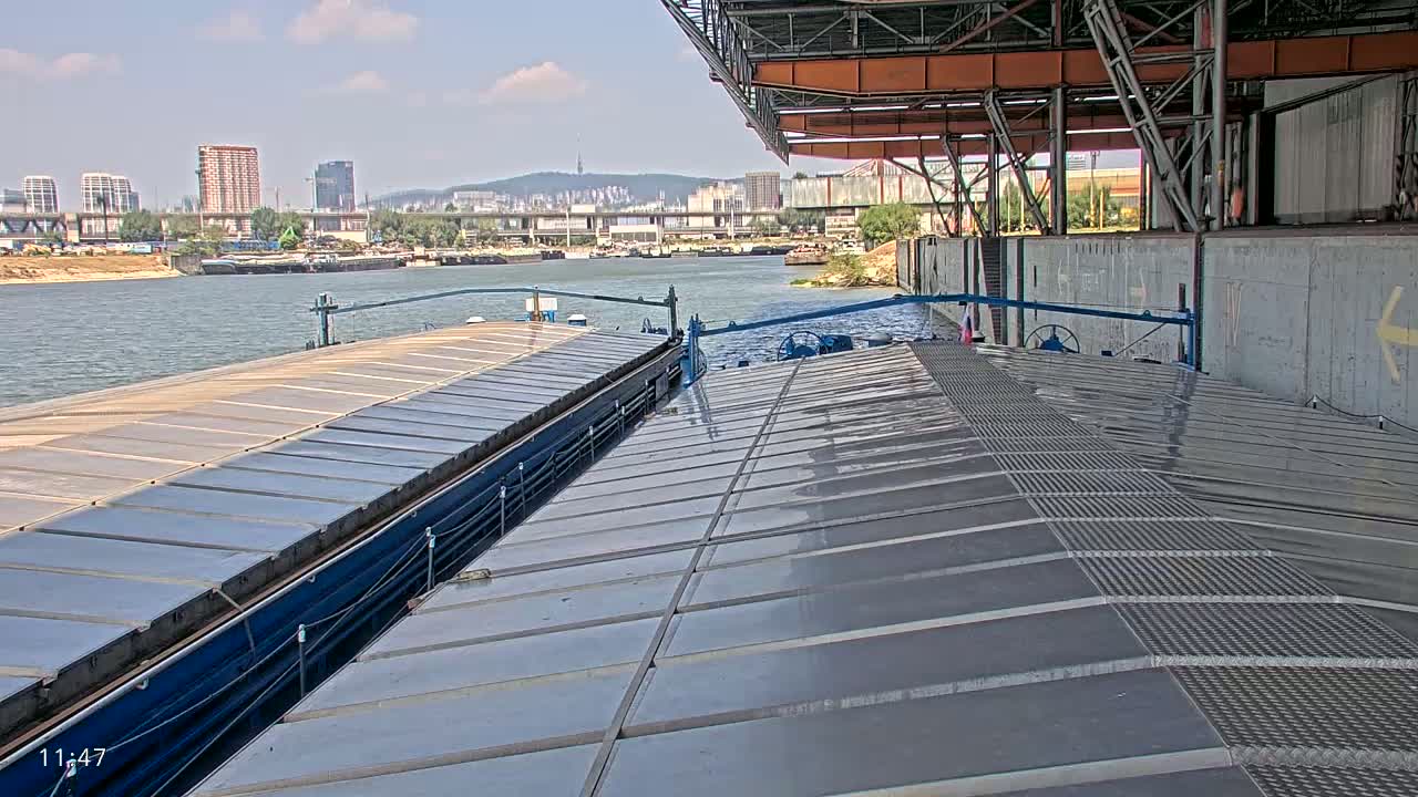 Two barges are docked at a concrete structure on a sunny day, with a cityscape and a river visible in the background.
