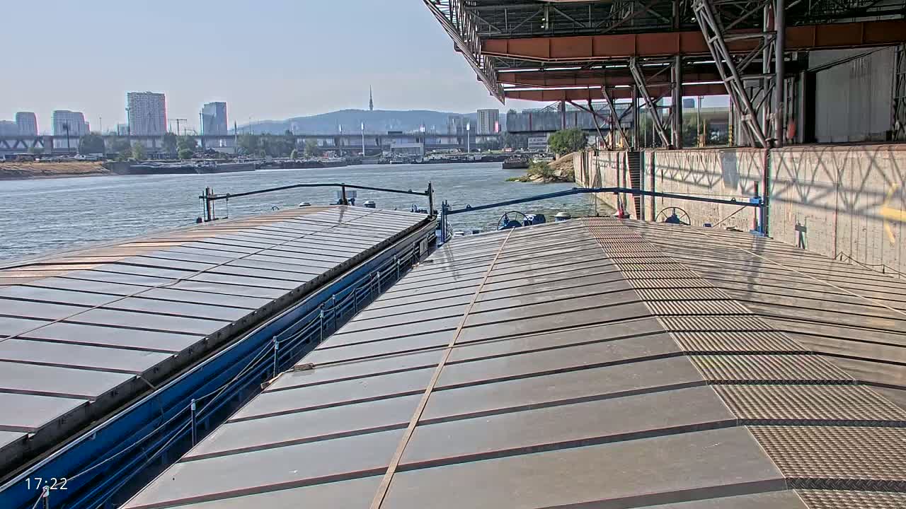 The deck of a barge is shown, with another barge visible in the background on a calm river under a sunny sky, and a city skyline in the distance.