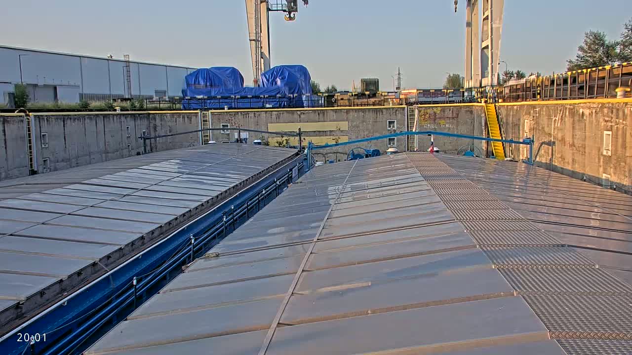 The flat, gray, paneled roof of a barge sits in a lock, with concrete walls and a crane visible in the background under a clear sky.