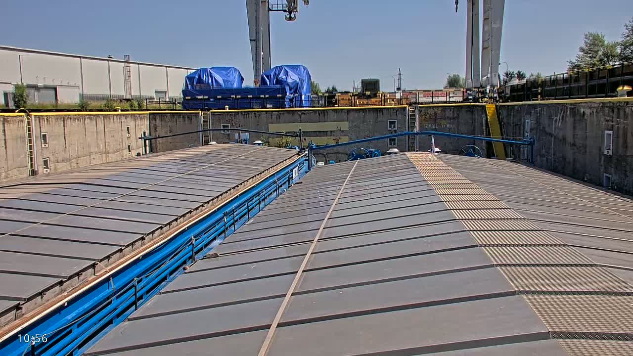 A view from the top of a barge shows a gray metal deck, flanked by blue metal structures, under a sunny sky.