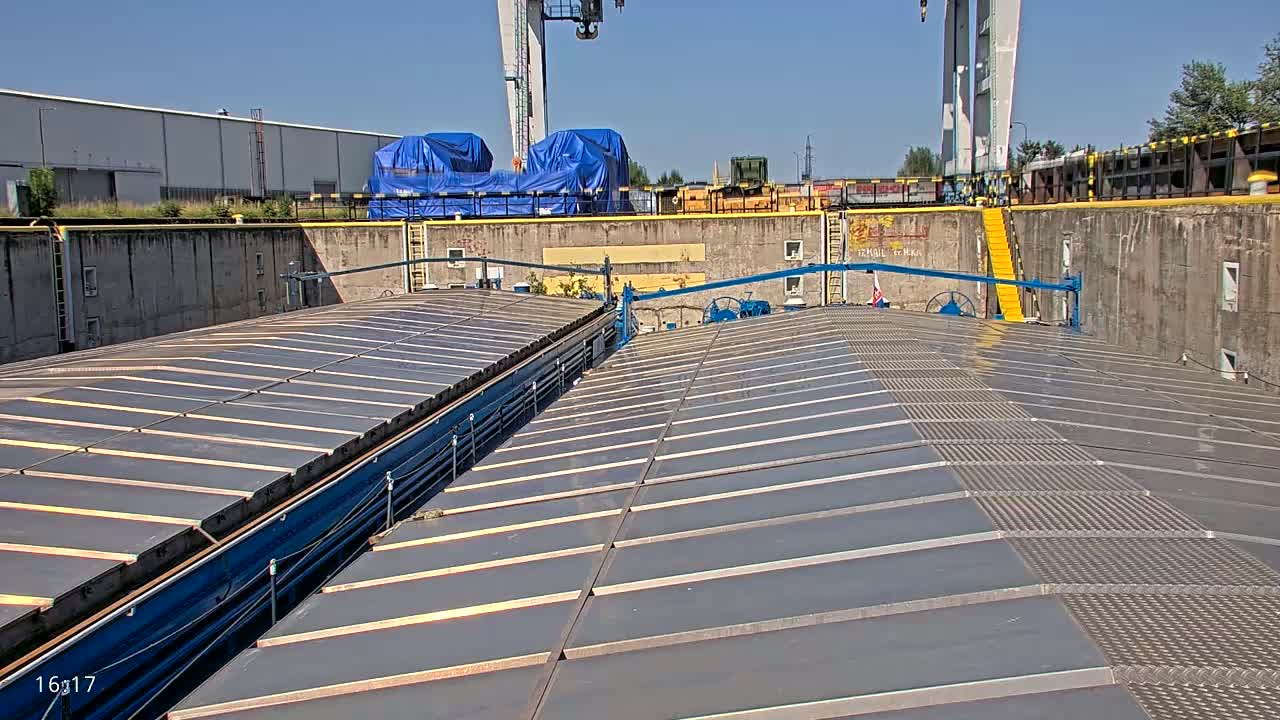 A flat, gray, sectioned deck of a barge sits in a lock, under a sunny sky.