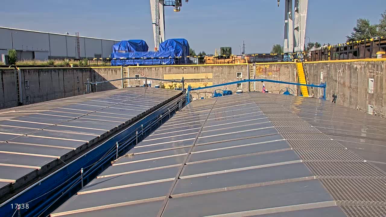 A view from atop a barge’s deck, showing a long, flat, gray surface divided into sections, under a sunny sky.